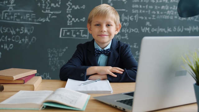 portrait of happy child wunderkind wearing suit sitting at desk in class with chalkboard full of formulas behind him. Education and knowledge concept